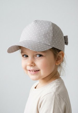Young child smiling, wearing a light gray polka dot baseball cap and a beige shirt, against a plain light background.