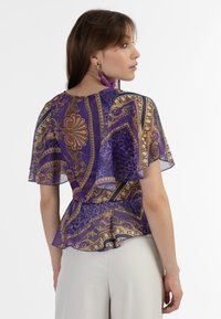 Fialová blůza se vzory a průhlednými, rozšířenými rukávy; má zlaté vzory a peplum v pase. Nosená s krémovými kalhotami.