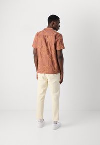 Camicetta a maniche corte color ruggine con ricami floreali, abbinata a pantaloni color crema e sneaker bianche, vista da dietro.