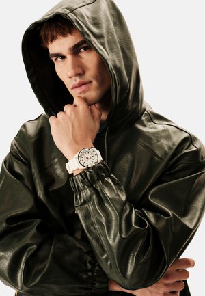 Grøn læderjakke med hætte og elastiske manchetter, kombineret med et hvidt ur med rund skive og sorte detaljer. Fokus på tekstur og materialer.