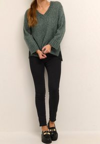 Pull en tricot vert avec un col en V, des poignets côtelés et des fentes sur les côtés, associé à un jean skinny noir et des mocassins noirs avec des accents dorés.