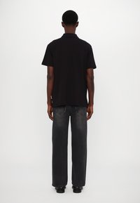 Svart polo-skjorte med myk tekstur og korte ermer, iført med løstsittende mørkegrå jeans og svarte sko, sett bakfra.