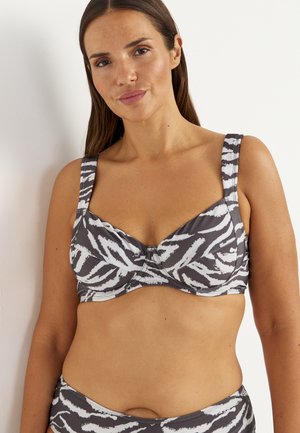 Woman with long brown hair wearing a black and white zebra-print bikini top, standing against a plain light background.