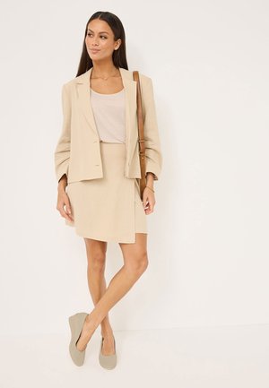 Beige skräddarsydd kavaj över en beige topp och en matchande omslagsskjorta, tillsammans med ljusgrå slip-on skor med en ren, slät textur.