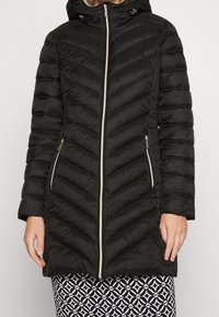 Schwarze Steppjacke mit Zickzacksteppung, Reißverschluss vorne und Kapuze. Mit goldenen Reißverschlüssen und Seitentaschen. Leichtes synthetisches Material.