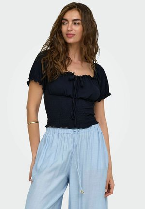 Femme aux cheveux bruns ondulés portant un haut noir froncé à manches courtes et un pantalon large bleu clair à cordon, regardant sur le côté.