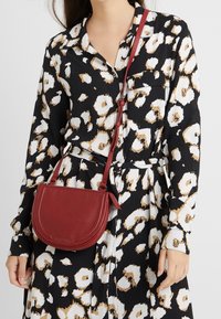 Czerwona skórzana torba na ramię o zakrzywionym kształcie, z teksturowaną powierzchnią i subtelnymi detalami przeszyć. Noszona z czarno-białą sukienką koszulową w wzory.