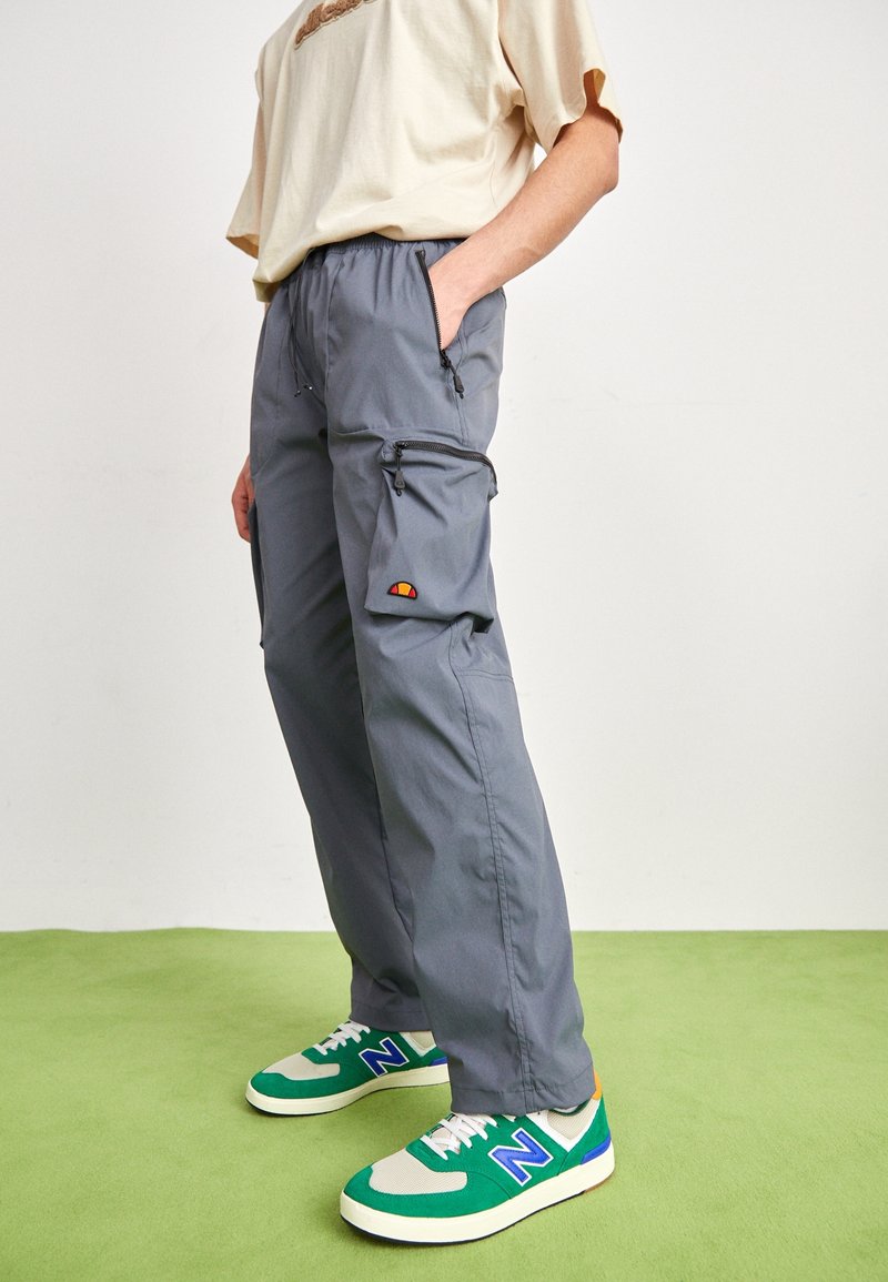 Personne portant un pantalon cargo gris avec des poches zippées et des baskets vertes New Balance debout sur un sol vert.