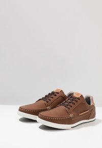 Paire de chaussures décontractées en cuir marron avec des lacets marron foncé, des accents de couture blancs et des semelles en caoutchouc blanc, posées sur une surface blanche.