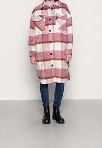 Rutig oversized skjortjacka i rosa och vitt med bruna accenter, med två bröstfickor och svarta knappar. Bärs med mörka jeans och stövlar.