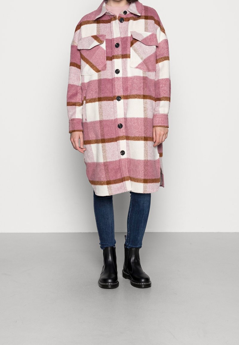 Rutig oversized skjortjacka i rosa och vitt med bruna accenter, med två bröstfickor och svarta knappar. Bärs med mörka jeans och stövlar.
