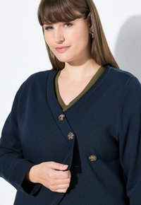 Donna con capelli castani lisci che indossa orecchini a cerchio dorati, un cardigan blu scuro con bottoni sopra una maglietta verde oliva a scollo a V.