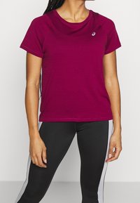 Camiseta deportiva color burdeos con mangas cortas, escote redondo, textura suave y pequeño logo en el pecho. Lleva puesta con leggings negros.