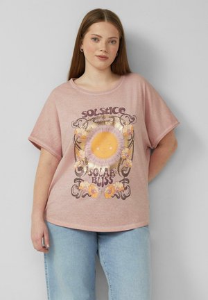 Jeune femme aux longs cheveux portant un t-shirt rose avec un motif soleil et fleurs portant l'inscription "Solstice Solar Bliss" et un jean bleu clair.
