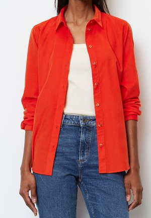 Person wearing a bright orange buttoned shirt over a white sleeveless top paired with high-waisted blue jeans, standing against a plain background.