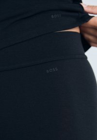 Detail zblízka na tmavú textíliu oblečenia s jemne embosovanými logami "BOSS" blízko pásu a spodného lemu, držaný rukou, ktorá zdvíha hornú vrstvu.