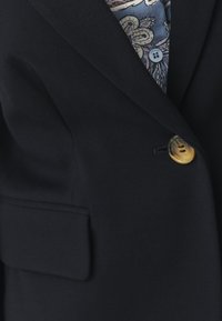 Blazer bleu marine en tissu texturé, doté d'une poche poitrine et d'un bouton doré. La doublure présente un motif cachemire en bleu et crème.