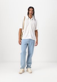 Chemise blanche à manches courtes avec des motifs géométriques, jean bleu clair et baskets beiges. Se marie avec un sac bandoulière de teinte neutre.