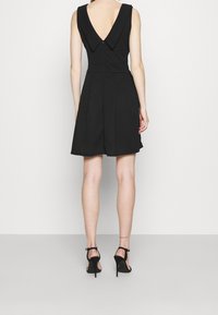 Žena v černých šatech bez rukávů s hlubokým V výstřihem na zádech a černých lodičkách na vysokém podpatku, stojí před jednoduchým světlým pozadím.