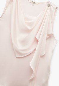 Lichtroze mouwloze blouse met gedrapeerde stof, gemaakt van een glad materiaal. Kenmerkt zich door een decoratieve zilveren accent bij de schoudernaad.