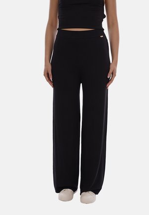 Donna indossa pantaloni neri a gamba larga abbinati a sneaker bianche e un top corto nero, in piedi davanti a uno sfondo semplice.