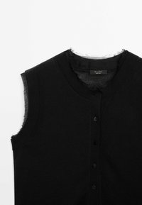 Haut noir sans manches à boutons avec bords bruts autour du col et des emmanchures sur fond blanc.