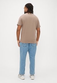 Ruskea puuvillainen t-paita, jossa on lyhyet hihat, nähtynä takaa, yhdistettynä vaaleansinisistä farkuista tehtyihin housuihin. Kangas näytt�ää pehmeältä ja rentoilta.