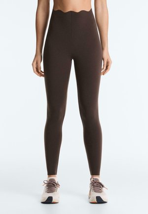 Nő magas derekú sötétbarna leggingsben, fodros derékszéllel, fehér és szürke sportcipőkkel, narancssárga díszítésekkel.