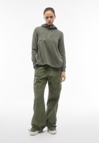 Felpa verde oliva con zip a metà e tasca a canguro, abbinata a pantaloni cargo verdi oliva a gamba larga con tasche laterali e riprese.