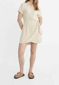 Vestito camicia beige a maniche corte con bottoni frontali, taschino sul petto e dettaglio con lacci laterali. Abbinato a sandali marroni con fibbie frontali.