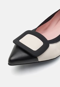 Sparktå sko med en ovandel i svart och beige läder, med en stor svart fyrkantig detalj på framsidan och en mjuk rosa inredning.