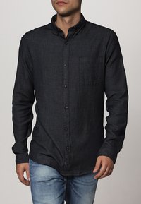Camicia button-up in denim scuro con colletto strutturato, taschino frontale e bottoni marroni, caratterizzata da un tessuto testurizzato e una vestibilità su misura.