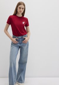 Red cotton t-shirt with a white graphic on the left chest, paired with light blue wide-leg denim jeans. Simple design, solid color.