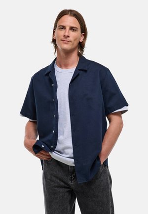 Jeune homme aux cheveux mi-longs portant une chemise bleue marine ouverte à manches courtes sur un t-shirt gris clair et un jean noir, les mains dans les poches.