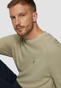 Beżowy sweter z dzianiny z okrągłym dekoltem, prążkowanymi mankietami i małą naszywką z logo na piersi. Noszony z ciemnymi spodniami, podkreślający fakturę i dopasowanie.