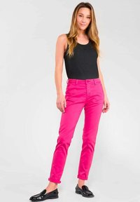 Eingefärbte rosa Hose aus glattem Stoff, mit einem Standardbund und Taschen, kombiniert mit einem schwarzen Trägershirt und schwarzen Loafers.