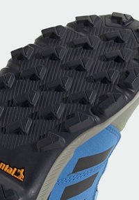 Niebieski but sportowy z teksturowaną szarą podeszwą zewnętrzną, wyposażony w agresywne wypustki dla lepszej przyczepności oraz wyraźne pomarańczowe logo marki po boku.