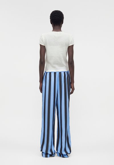 Άτομο όρθιο με την πλάτη στραμμένη, φοράει λευκή ριμπ μπλούζα με κοντά μανίκια και φαρδιά παντελόνια με κάθετες μπλε και μαύρες ρίγες.