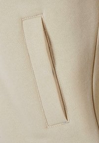 Gros plan sur un tissu beige montrant une poche passepoilée rectangulaire avec des coutures soignées sur un textile à texture lisse.