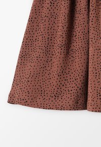 La tela marrón presenta pequeños lunares negros. El material parece suave con un ligero brillo, terminado con un dobladillo recto y caída casual.