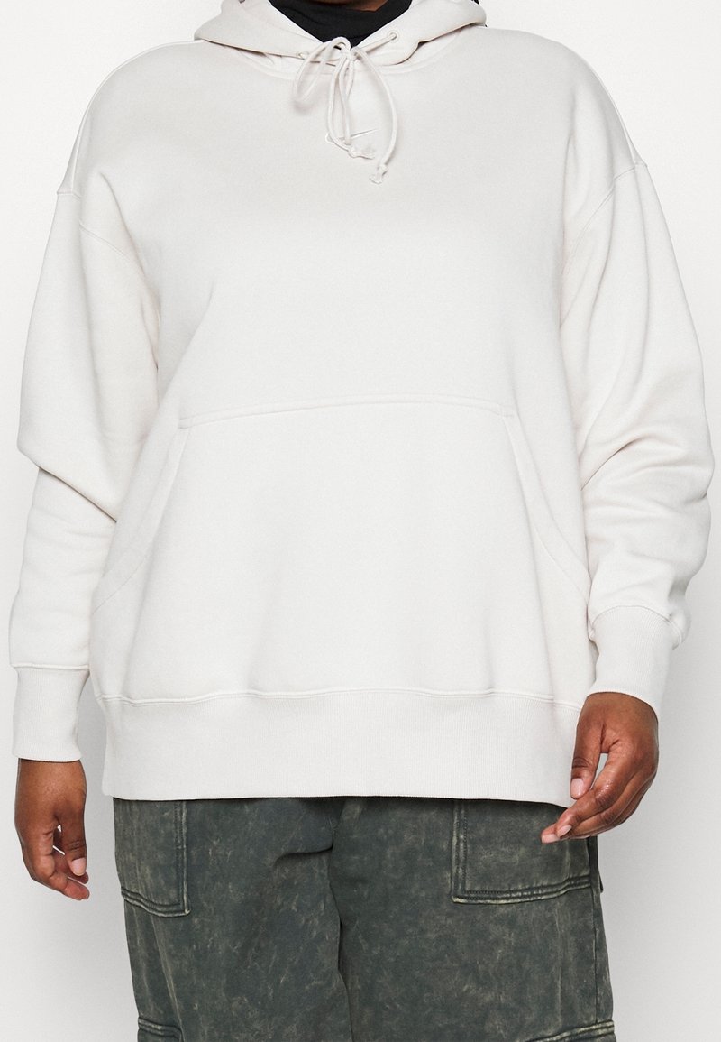 Přes veliká krémová mikina s kapucí, šňůrkou, přední kapsou a žebrovanými manžetami, spojená s tmavě zelenými cargo kalhotami s mnoha kapsami.