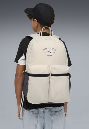 Boy wearing black cap and black T-shirt carries beige Puma Athletics backpack with two front zip pockets and mesh side pockets.
