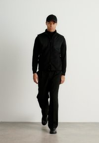 Colete preto com fecho de correr à frente por cima de uma camisola preta de gola alta, combinado com calças pretas e sapatos, usado por uma pessoa a caminhar contra um fundo simples.