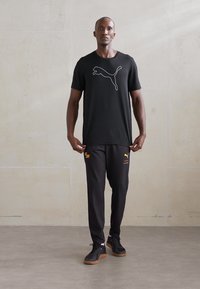 Crna majica kratkih rukava s bijelim PUMA logotipom, uparena s crnim hlačama s žutim detaljima i PUMA logotipom. Obuća je crna s gumiranim potplatom.