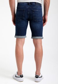 Pantalones cortos de mezclilla azul oscuro con dobladillos doblados, que cuentan con bolsillos traseros y una textura suave. Llevados con zapatillas blancas.