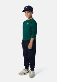 Grüner Sweatshirt mit rundem Logo, marineblaue Jogginghose und weiße Sneakers mit beigen Akzenten. Das Kind trägt eine passende marineblaue Mütze.