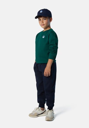 Felpa verde con un logo rotondo, pantaloni della tuta blu navy e sneakers bianche con dettagli beige. Il bambino indossa un berretto blu navy abbinato.