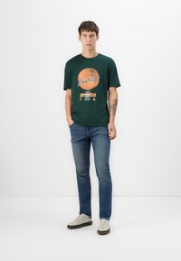Mørkegrøn bomulds t-shirt med et cirkulært grafik, hvor der står "Endless" og "arena" i orange. Kombineret med blå denim jeans og lyse sneakers.