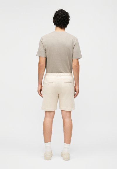 Άνδρας με σγουρά μαλλιά στέκεται με την πλάτη γυρισμένη, φοράει μπεζ πουκάμισο με κοντά μανίκια, εκρού σορτς, λευκές κάλτσες και μπεζ αθλητικά σε λευκό φόντο.