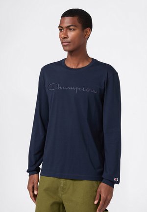 Jonge man in een marineblauwe langekous shirt met het "Champion" logo geborduurd op de borst en olijfgroene broek, staande tegen een witte achtergrond.